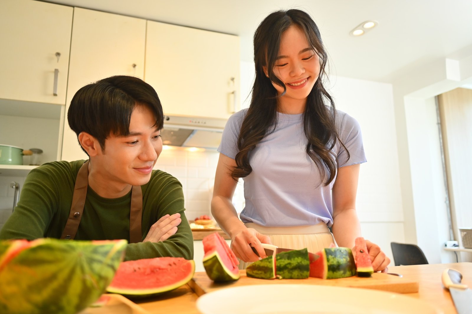 Pretty young woman and boyfriend cutting watermelon on wooden kitchen tabletop
